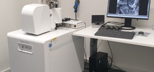 Ein Rasterelektronenmikroskop steht in einem hellen, klinisch wirkenden Raum. Das Gerät ist weiß, quaderförmig und besitzt einen zylindrischen Aufsatz mit mehreren metallischen Komponenten. Daneben befindet sich ein Schreibtisch mit einem Computer, auf dessen Monitor eine schwarz-weiße Nahaufnahme einer mikroskopischen Struktur angezeigt wird. Tastatur, Maus und weitere technische Geräte liegen auf dem Tisch. Die Umgebung wirkt aufgeräumt und funktional.