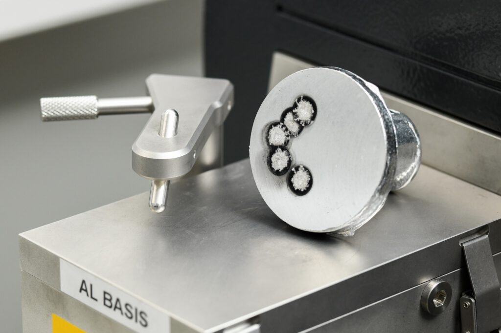 Nahaufnahme eines zylindrischen Probenstücks aus einer Aluminiumlegierung, das auf einer metallischen Oberfläche mit der Aufschrift 'AL BASIS' liegt. Die Stirnfläche der Probe zeigt mehrere kreisförmige Vertiefungen mit dunklem Rand und hellem Zentrum, die auf eine Analyse der Schmelze, vermutlich mittels optischer Emissionsspektrometrie (OES), hinweisen. Links daneben befindet sich ein metallischer Haltearm mit Rändelschraube. Die Szene ist technisch und klar ausgeleuchtet.