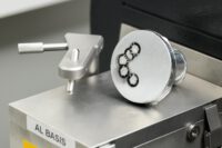 Nahaufnahme eines zylindrischen Probenstücks aus einer Aluminiumlegierung, das auf einer metallischen Oberfläche mit der Aufschrift 'AL BASIS' liegt. Die Stirnfläche der Probe zeigt mehrere kreisförmige Vertiefungen mit dunklem Rand und hellem Zentrum, die auf eine Analyse der Schmelze, vermutlich mittels optischer Emissionsspektrometrie (OES), hinweisen. Links daneben befindet sich ein metallischer Haltearm mit Rändelschraube. Die Szene ist technisch und klar ausgeleuchtet.