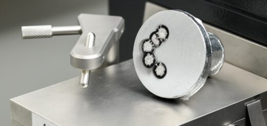 Nahaufnahme eines zylindrischen Probenstücks aus einer Aluminiumlegierung, das auf einer metallischen Oberfläche mit der Aufschrift 'AL BASIS' liegt. Die Stirnfläche der Probe zeigt mehrere kreisförmige Vertiefungen mit dunklem Rand und hellem Zentrum, die auf eine Analyse der Schmelze, vermutlich mittels optischer Emissionsspektrometrie (OES), hinweisen. Links daneben befindet sich ein metallischer Haltearm mit Rändelschraube. Die Szene ist technisch und klar ausgeleuchtet.