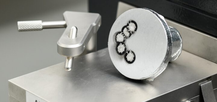 Nahaufnahme eines zylindrischen Probenstücks aus einer Aluminiumlegierung, das auf einer metallischen Oberfläche mit der Aufschrift 'AL BASIS' liegt. Die Stirnfläche der Probe zeigt mehrere kreisförmige Vertiefungen mit dunklem Rand und hellem Zentrum, die auf eine Analyse der Schmelze, vermutlich mittels optischer Emissionsspektrometrie (OES), hinweisen. Links daneben befindet sich ein metallischer Haltearm mit Rändelschraube. Die Szene ist technisch und klar ausgeleuchtet.