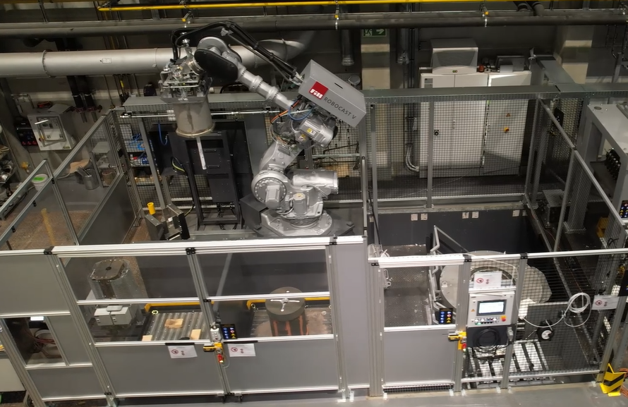 Industrieroboterarm des Gießroboters RoboCast V in einer automatisierten Fertigungszelle, umgeben von Metallgittern und Schutzwänden. Der Roboterarm ist zentral positioniert und mit einem Injektor ausgestattet. Im Hintergrund sind technische Anlagen, Rohre und weitere Maschinenteile sichtbar. Ein Bedienfeld mit Display und Bedienelementen befindet sich rechts im Vordergrund. Die Umgebung wirkt sauber und strukturiert.